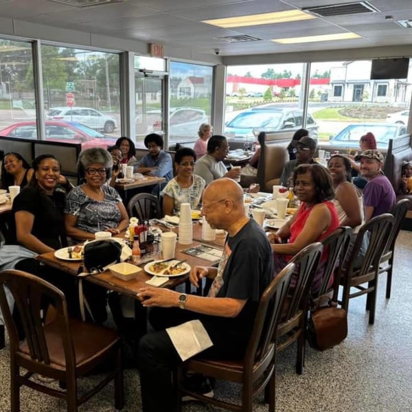Diverse group of people enjoying a meal together at Chef Flavas Restaurant, with large windows overlooking a street.