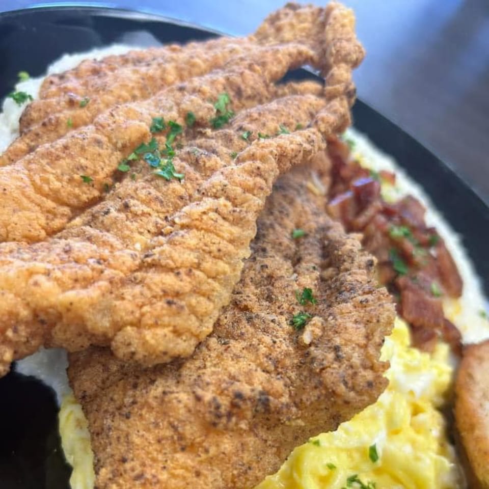 Chef Flavas' soul-food dish featuring crispy fried fish, creamy grits, scrambled eggs, and bacon.
