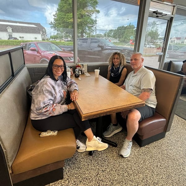 Three people smiling at a booth table inside Chef Flavas Restaurant with large windows overlooking a street.