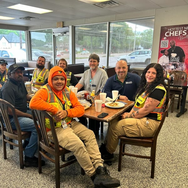 Seven people in work attire, some wearing high-visibility vests, smiling at a table in Chef Flavas Restaurant.